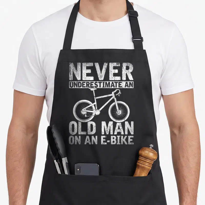 Funny underestimate bicycle apron - White e-bike centered between distressed text reading Never Underestimate An Old — shown on a black cotton canvas apron against a white background