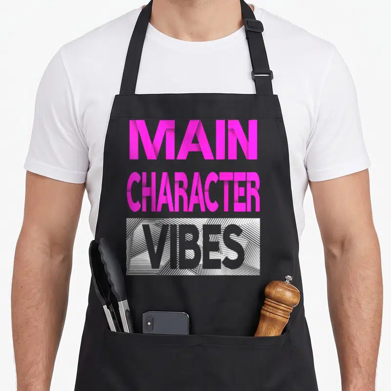 Cute main character aprons - Stacked pink and grey text reading MAIN CHARACTER VIBES with diagonal lined rectangle — shown on a black cotton canvas apron against a white background