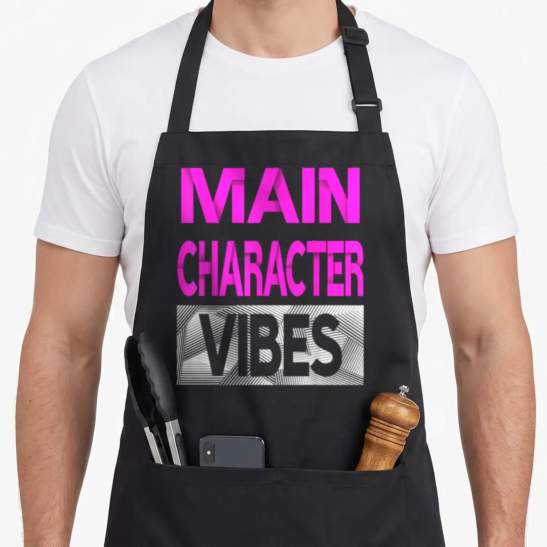 Cute main character aprons - Stacked pink and grey text reading MAIN CHARACTER VIBES with diagonal lined rectangle — shown on a black cotton canvas apron against a white background