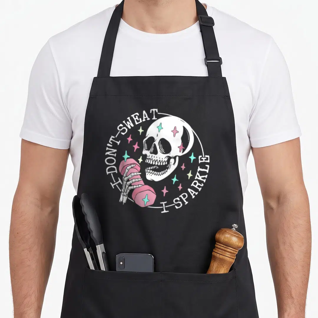 Sweat skeleton apron - Cartoon skull holding pink dumbbell surrounded by sparkles with I Don't Sweat I Sparkle text — shown on a black cotton canvas apron against a white background