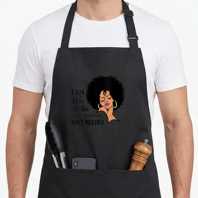 Sassy african print apron - Cartoon woman with black afro and yellow hoops beside stacked text I AM Who I Am YOUR — shown on a black cotton canvas apron against a white background