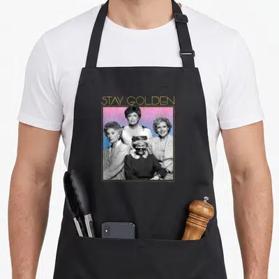 Cute stay golden apron - STAY GOLDEN text above four women on pink blue halftone dot background — shown on a black cotton canvas apron against a white background