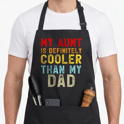 Definitely favorite aunt apron - Stacked colorful text reading MY AUNT IS DEFINITELY COOLER THAN MY DAD in red orange — shown on a black cotton canvas apron against a white background