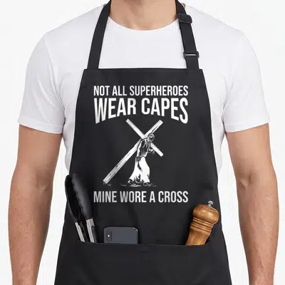 Superhero cross apron - White text and line art of Jesus carrying a cross on a black background — shown on a black cotton canvas apron against a white background