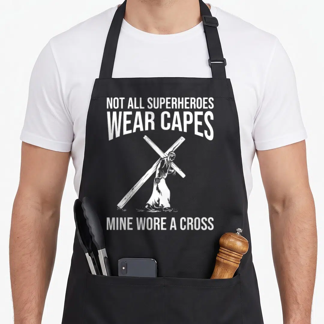 Superhero cross apron - White text and line art of Jesus carrying a cross on a black background — shown on a black cotton canvas apron against a white background
