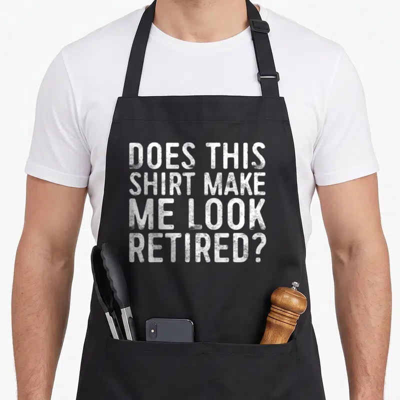 Doe retirement apron - White distressed text reading DOES THIS SHIRT MAKE ME LOOK RETIRED stacked on black fabric — shown on a black cotton canvas apron against a white background