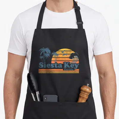 Siesta spring aprons - Retro blue and orange striped sun with palm trees birds wave and Siesta Key Florida text — shown on a black cotton canvas apron against a white background