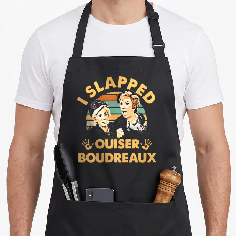 Slapped comfort apron - I Slapped Ouiser Boudreaux text with two women portraits and handprints on retro striped sun — shown on a black cotton canvas apron against a white background