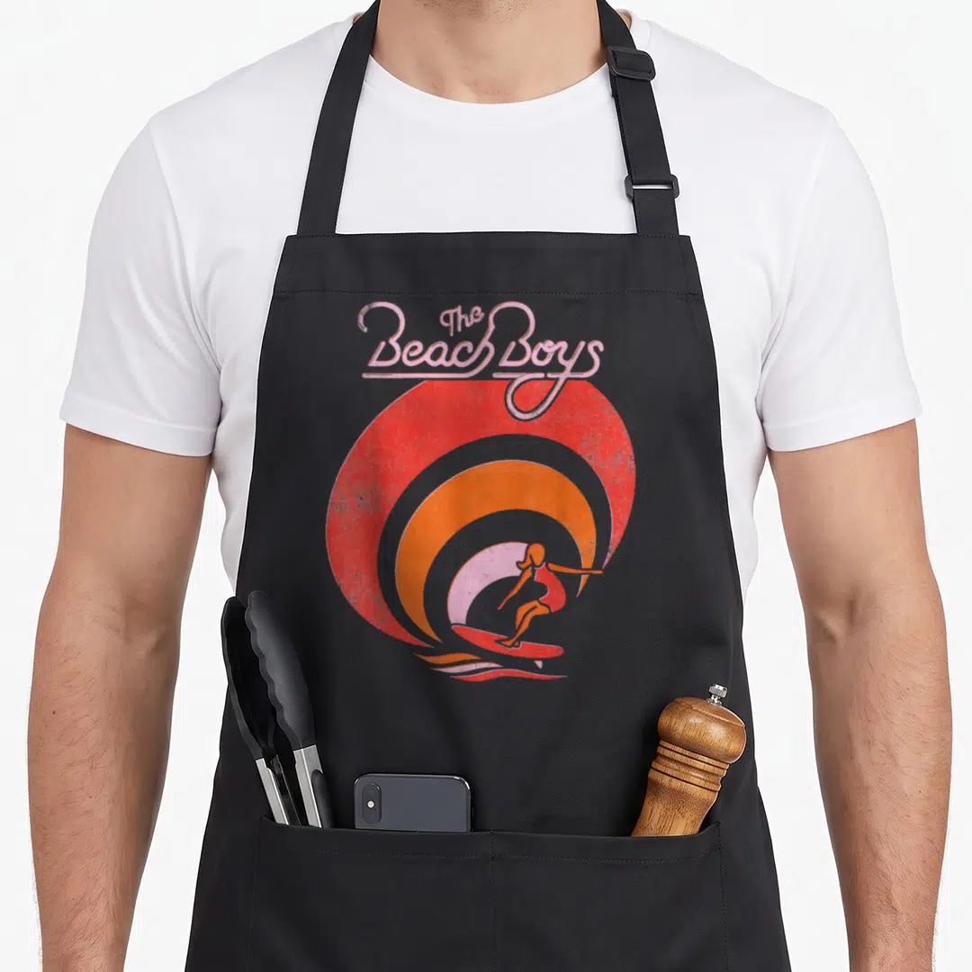 Beach surfer apron - The Beach Boys text over red orange pink wave with surfer silhouette — shown on a black cotton canvas apron against a white background