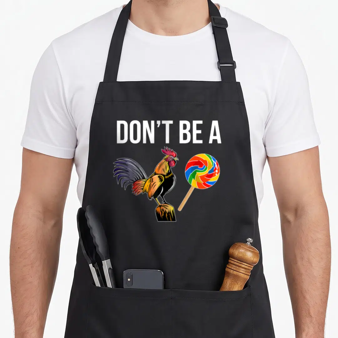 Sei rooster apron - DON'T BE A text above a rooster on a post beside a rainbow swirl lollipop — shown on a black cotton canvas apron against a white background