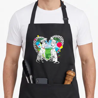 Pongo perdita dalmatian apron - Four dalmatians inside a spotted heart with balloons and green landscape background — shown on a black cotton canvas apron against a white background