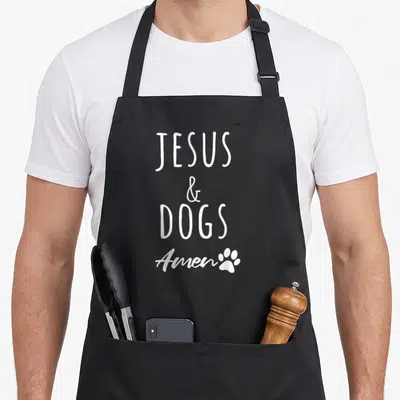 Christ dog apron - White stacked text JESUS & DOGS Amen with a small paw print on a black background — shown on a black cotton canvas apron against a white background