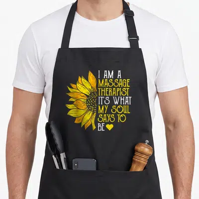 Sunflower massage therapist apron - Yellow sunflower graphic beside white and yellow stacked text ending with a small — shown on a black cotton canvas apron against a white background