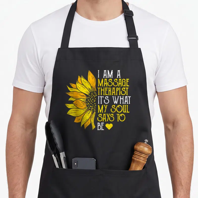 Sunflower massage therapist apron - Yellow sunflower graphic beside white and yellow stacked text ending with a small — shown on a black cotton canvas apron against a white background