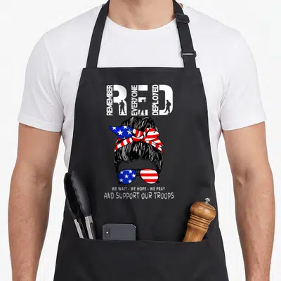 Friday support military apron - Woman's head with patriotic bun and sunglasses under REMEMBER RED EVERYONE DEPLOYED text — shown on a black cotton canvas apron against a white background