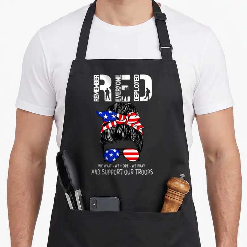 Friday support military apron - Woman's head with patriotic bun and sunglasses under REMEMBER RED EVERYONE DEPLOYED text — shown on a black cotton canvas apron against a white background