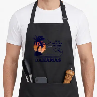 Better bahamian apron - Retro sunset graphic with palm trees, seagulls, and wave in dark blue and orange — shown on a black cotton canvas apron against a white background