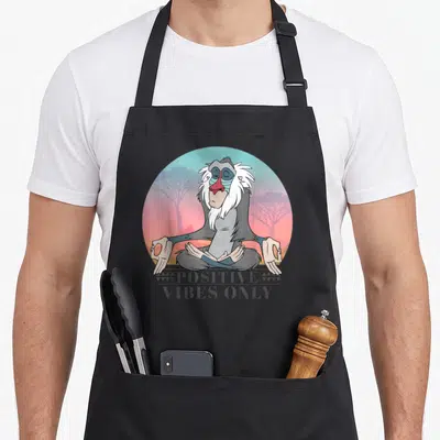 Rafiki positive vibes apron - Mandrill meditating in a lotus position within a sunset circle with stacked text below — shown on a black cotton canvas apron against a white background