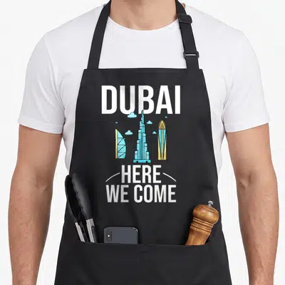 Uae travel aprons - DUBAI text, three skyscrapers, HERE WE COME, white curves, light blue clouds — shown on a black cotton canvas apron against a white background
