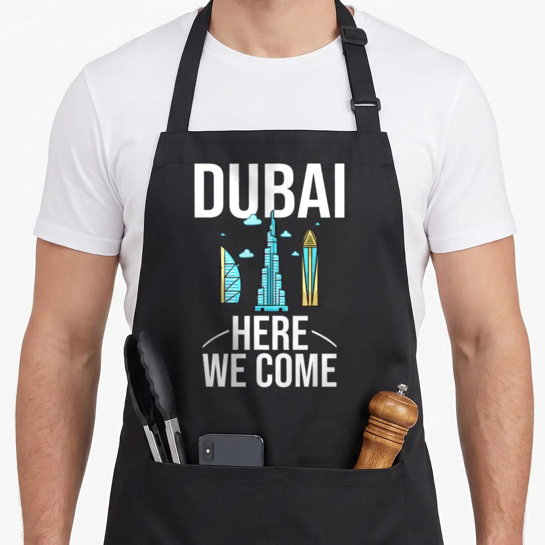 Uae travel aprons - DUBAI text, three skyscrapers, HERE WE COME, white curves, light blue clouds — shown on a black cotton canvas apron against a white background