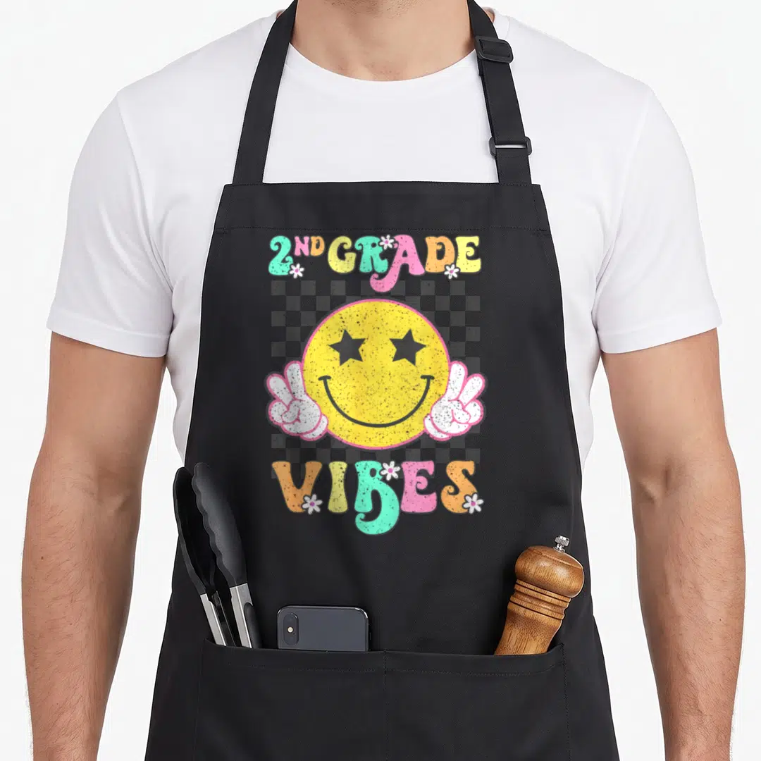 Smile 2nd apron - Retro yellow smiley face with star eyes and peace signs surrounded by 2ND GRADE VIBES text — shown on a black cotton canvas apron against a white background