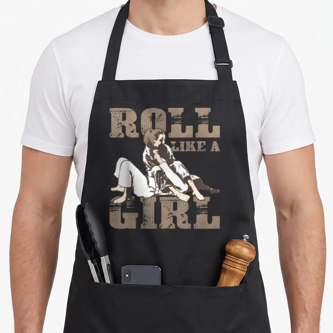 Roll brazilian apron - Distressed tan ROLL LIKE A GIRL text surrounding a woman in white gi grappling an opponent — shown on a black cotton canvas apron against a white background