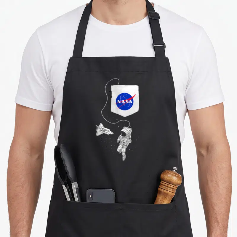 Faux nasa apron - Printed white pocket with NASA logo and floating astronaut shuttle cartoon — shown on a black cotton canvas apron against a white background