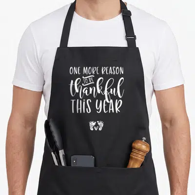 Reason thankful apron - white text ONE MORE REASON TO BE thankful THIS YEAR above pair of baby footprints with heart — shown on a black cotton canvas apron against a white background