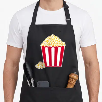 Retro popcorn apron - Cartoon red and white striped bucket filled with yellow and white popcorn with loose cluster — shown on a black cotton canvas apron against a white background
