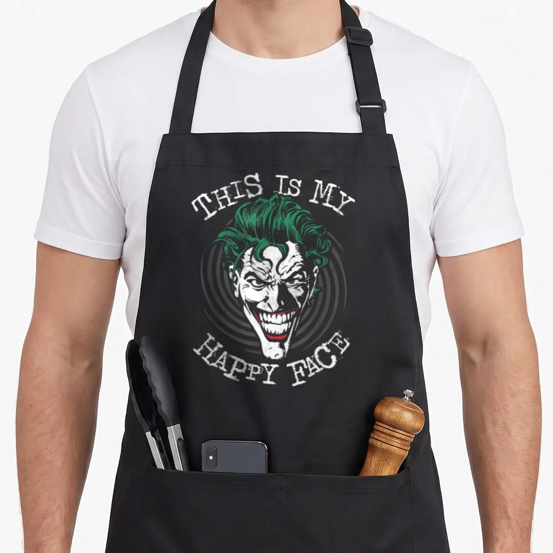 Retro face apron - The Joker head with green hair and red smile over grey spiral with arched text — shown on a black cotton canvas apron against a white background