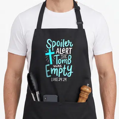 Alert easter apron - Vertical text Spoiler Alert The Tomb Was Empty Luke 24:24 with light blue cross and white rays — shown on a black cotton canvas apron against a white background