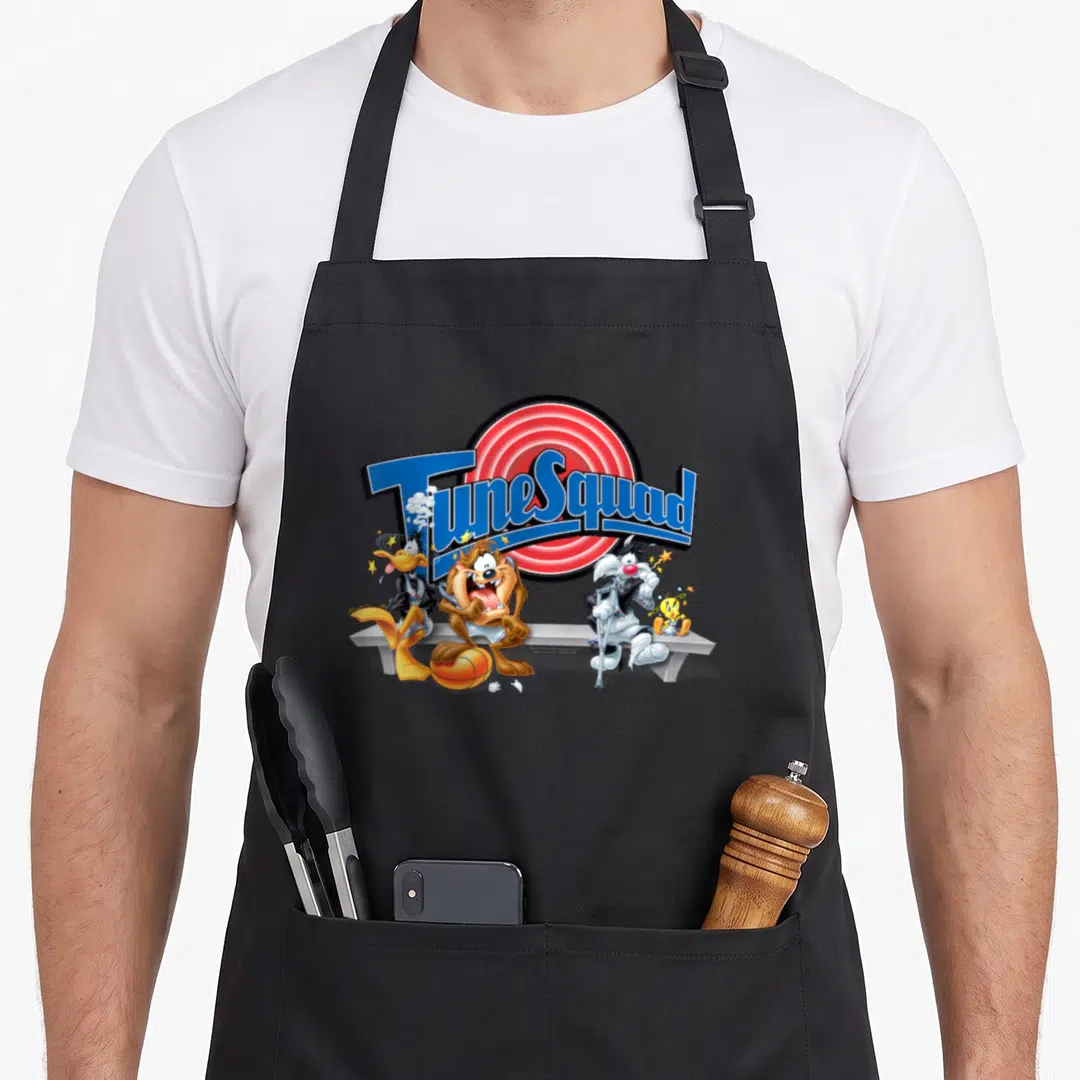 Space k squad apron - TuneSquad text over red circle with four cartoon characters on a bench and an orange basketball — shown on a black cotton canvas apron against a white background