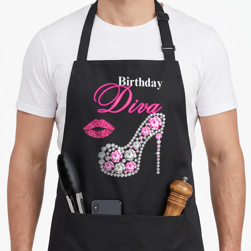 Heel hot pink apron - Birthday Diva text above pink lips and a gem-encrusted high heel shoe — shown on a black cotton canvas apron against a white background