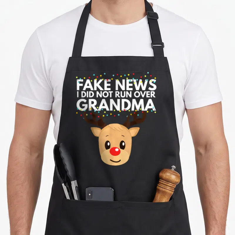 Casual holiday christmas aprons - Cartoon reindeer head with white text FAKE NEWS I DID NOT RUN OVER GRANDMA — shown on a black cotton canvas apron against a white background