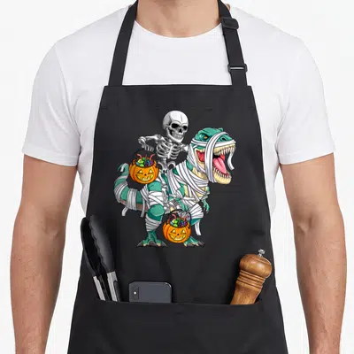 Skeleton riding halloween apron - Cartoon mummy T-Rex and skeleton rider holding two orange pumpkin candy buckets — shown on a black cotton canvas apron against a white background