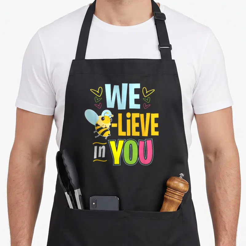 Test bee aprons - Stacked WE -LIEVE In YOU text with cartoon bee and three hearts in blue yellow green pink — shown on a black cotton canvas apron against a white background