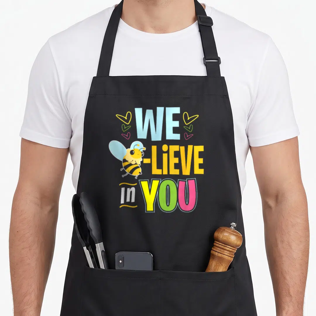 Test bee aprons - Stacked WE -LIEVE In YOU text with cartoon bee and three hearts in blue yellow green pink — shown on a black cotton canvas apron against a white background