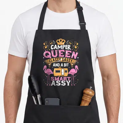 Classy smart apron - Stacked CAMPER QUEEN text with crown, orange camper, two pink flamingos, and decorative dots — shown on a black cotton canvas apron against a white background