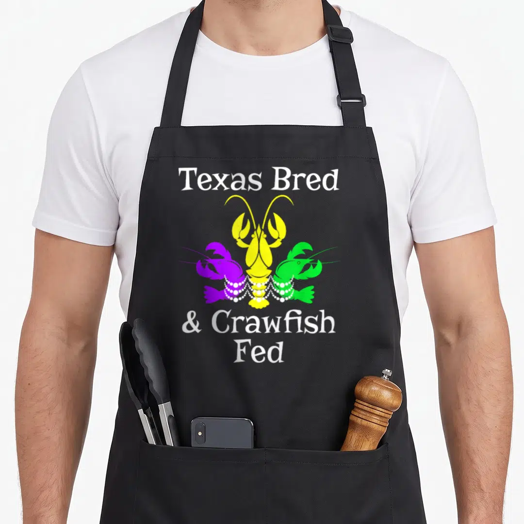 Texas fat chef apron - Three cartoon crawfish in purple yellow green wearing white pearl necklaces with Texas Bred text — shown on a black cotton canvas apron against a white background