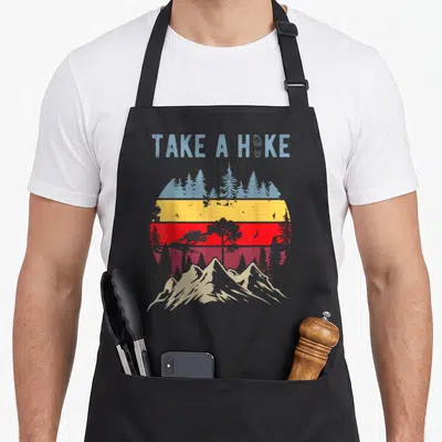 Hiking nature outdoor apron - Take a Hike text with footprint over distressed circular mountain landscape in red yellow — shown on a black cotton canvas apron against a white background