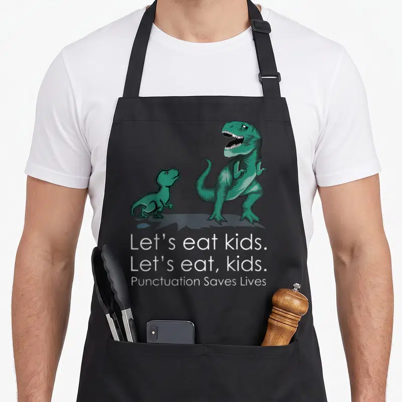 Live let's eat apron - Teal cartoon dinosaurs above white text reading Let's eat kids. and Punctuation Saves Lives — shown on a black cotton canvas apron against a white background