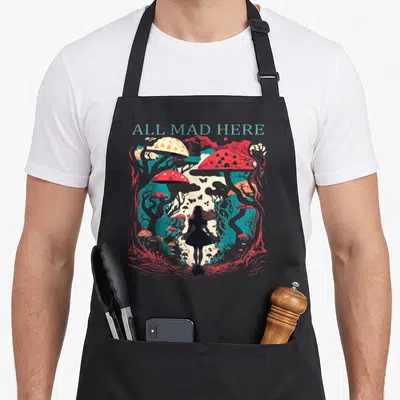 Cute mad alice wonderland apron - ALL MAD HERE text above girl silhouette in surreal forest with red mushrooms and teal — shown on a black cotton canvas apron against a white background