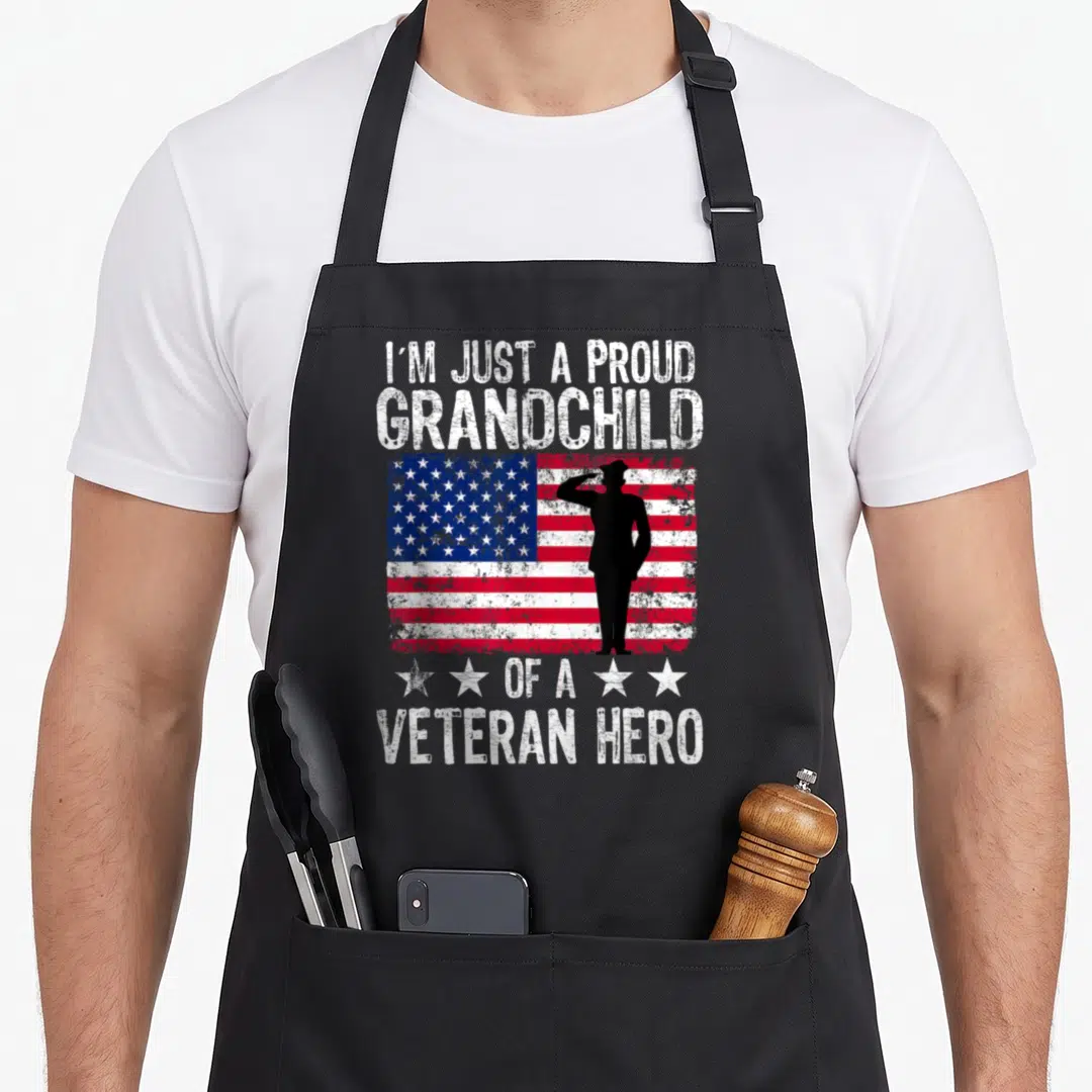 Grandchild american flag apron - Distressed flag with saluting soldier silhouette and proud grandchild veteran hero text — shown on a black cotton canvas apron against a white background