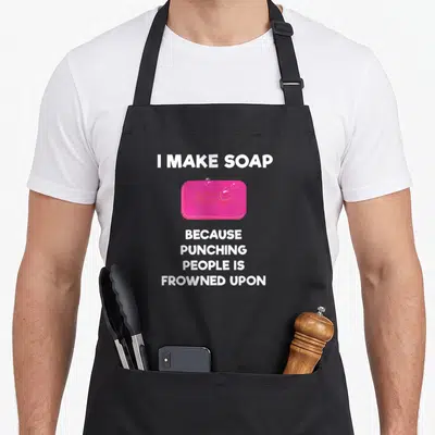 Soap maker apron - Pink cartoon soap bar with white text I make soap because punching people is frowned upon — shown on a black cotton canvas apron against a white background