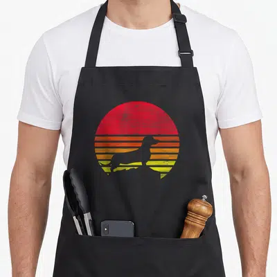 Funny dachshund apron - Grey dachshund silhouette against red orange and yellow retro striped circle — shown on a black cotton canvas apron against a white background