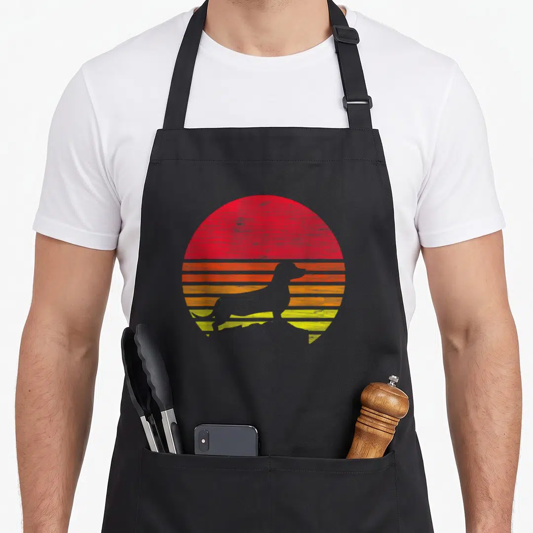 Funny dachshund apron - Grey dachshund silhouette against red orange and yellow retro striped circle — shown on a black cotton canvas apron against a white background