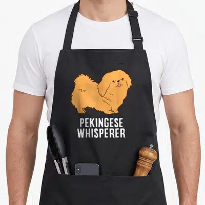 Pekingese whisperer dog apron - Cartoon tan Pekingese dog with pink tongue above white PEKINGESE WHISPERER text — shown on a black cotton canvas apron against a white background