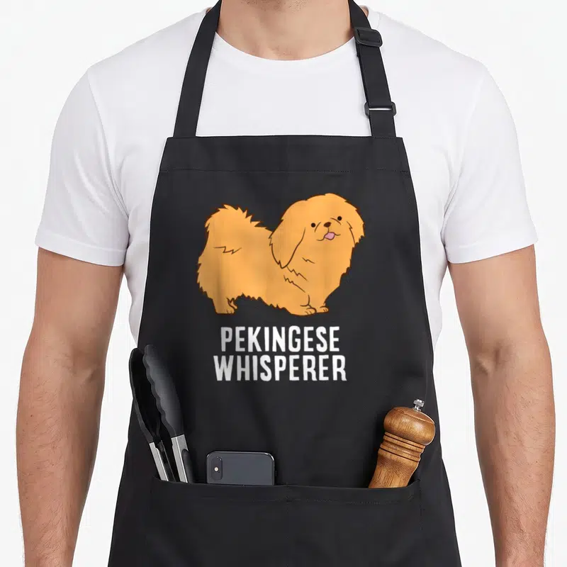 Pekingese whisperer dog apron - Cartoon tan Pekingese dog with pink tongue above white PEKINGESE WHISPERER text — shown on a black cotton canvas apron against a white background