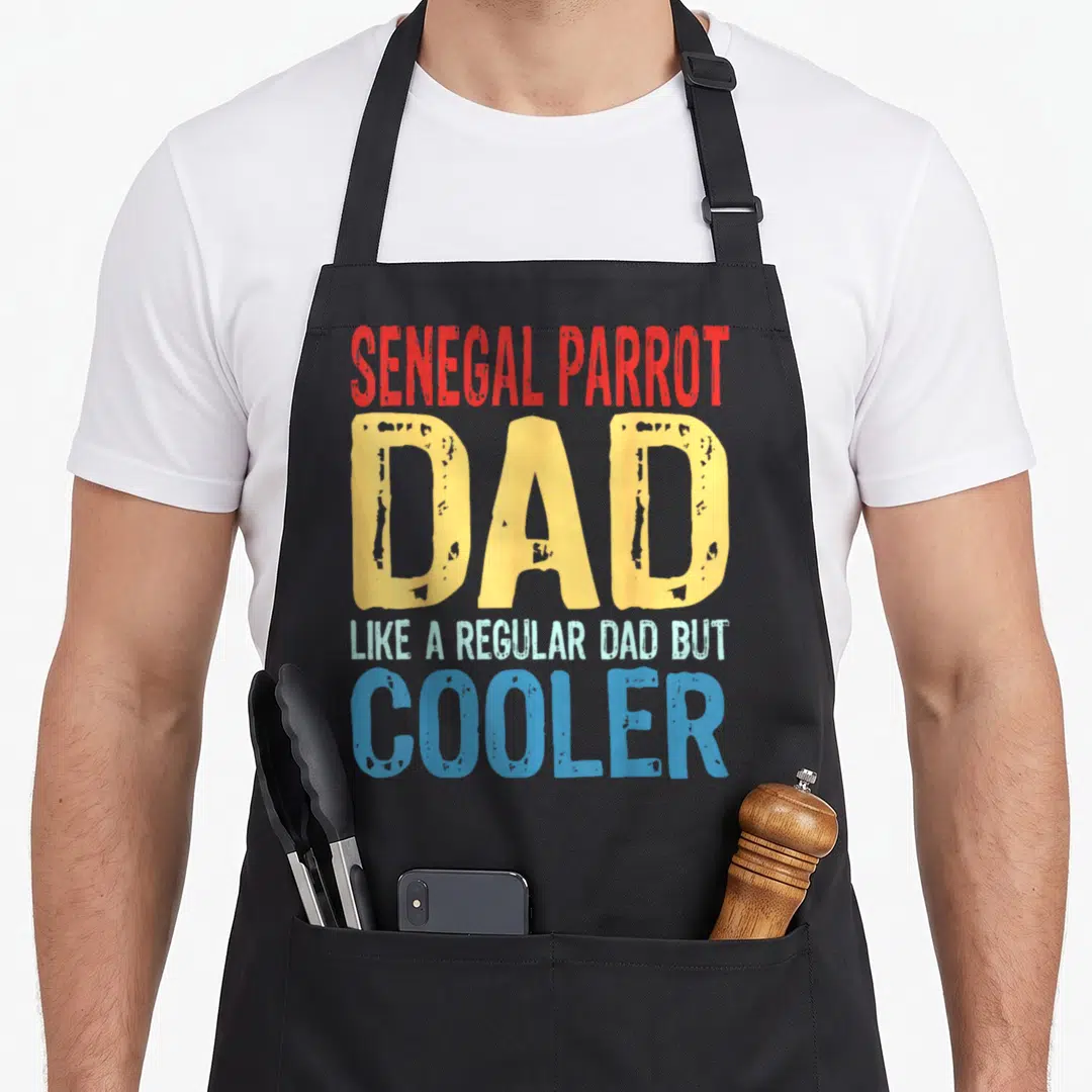 Senegal parrot dad apron - Red yellow and blue stacked text reading Senegal Parrot Dad Like A Regular Dad But Cooler — shown on a black cotton canvas apron against a white background
