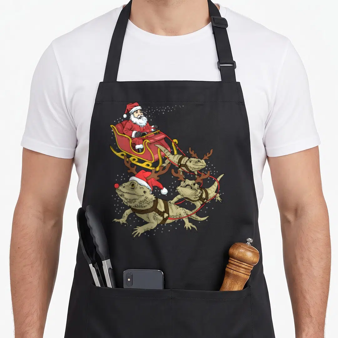Dragon christmas apron - Cartoon Santa sleigh pulled by three bearded dragons with antlers and red nose — shown on a black cotton canvas apron against a white background